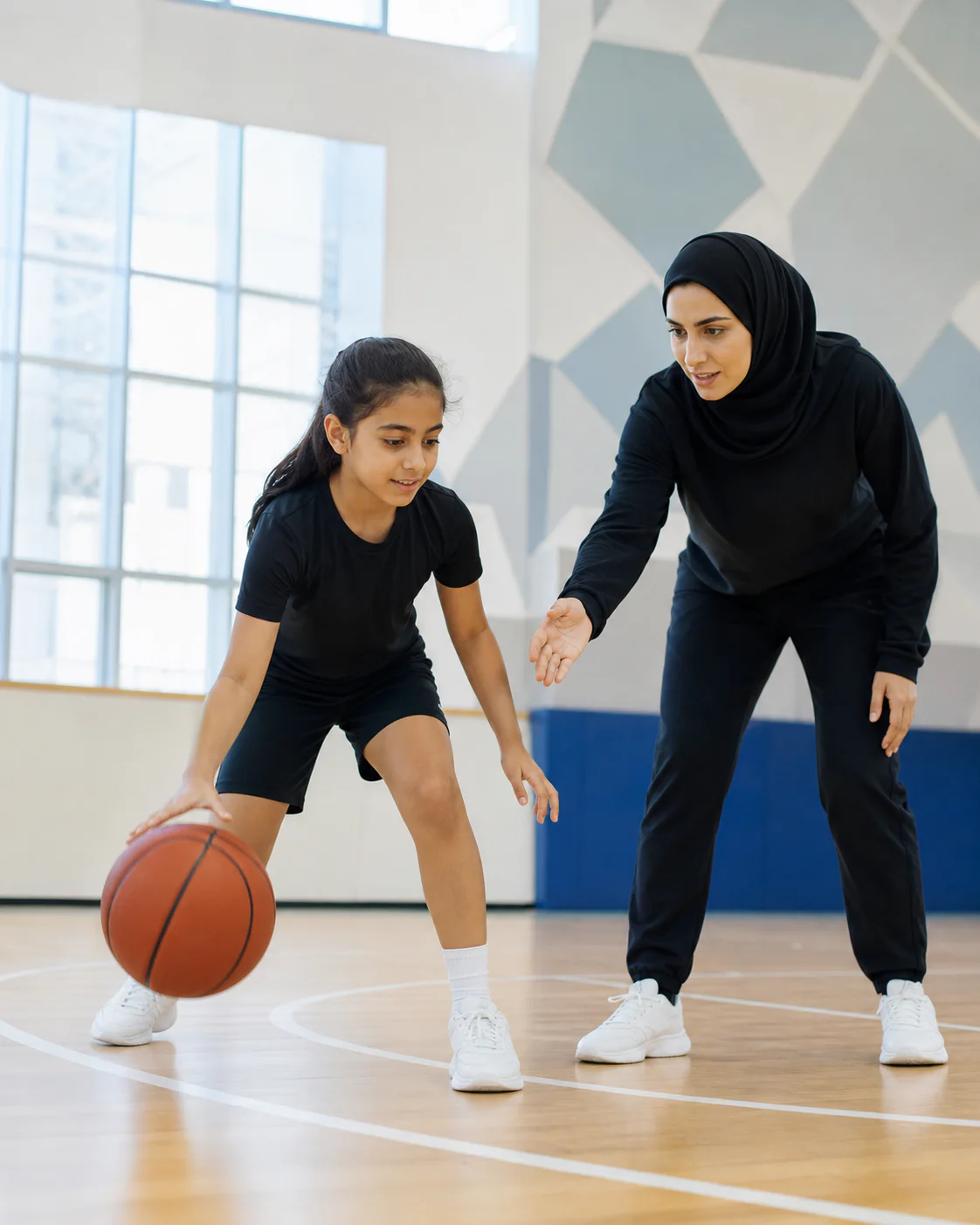 Young girl learning a controlled basketball dribble with her coach in a Dubai school sports hall