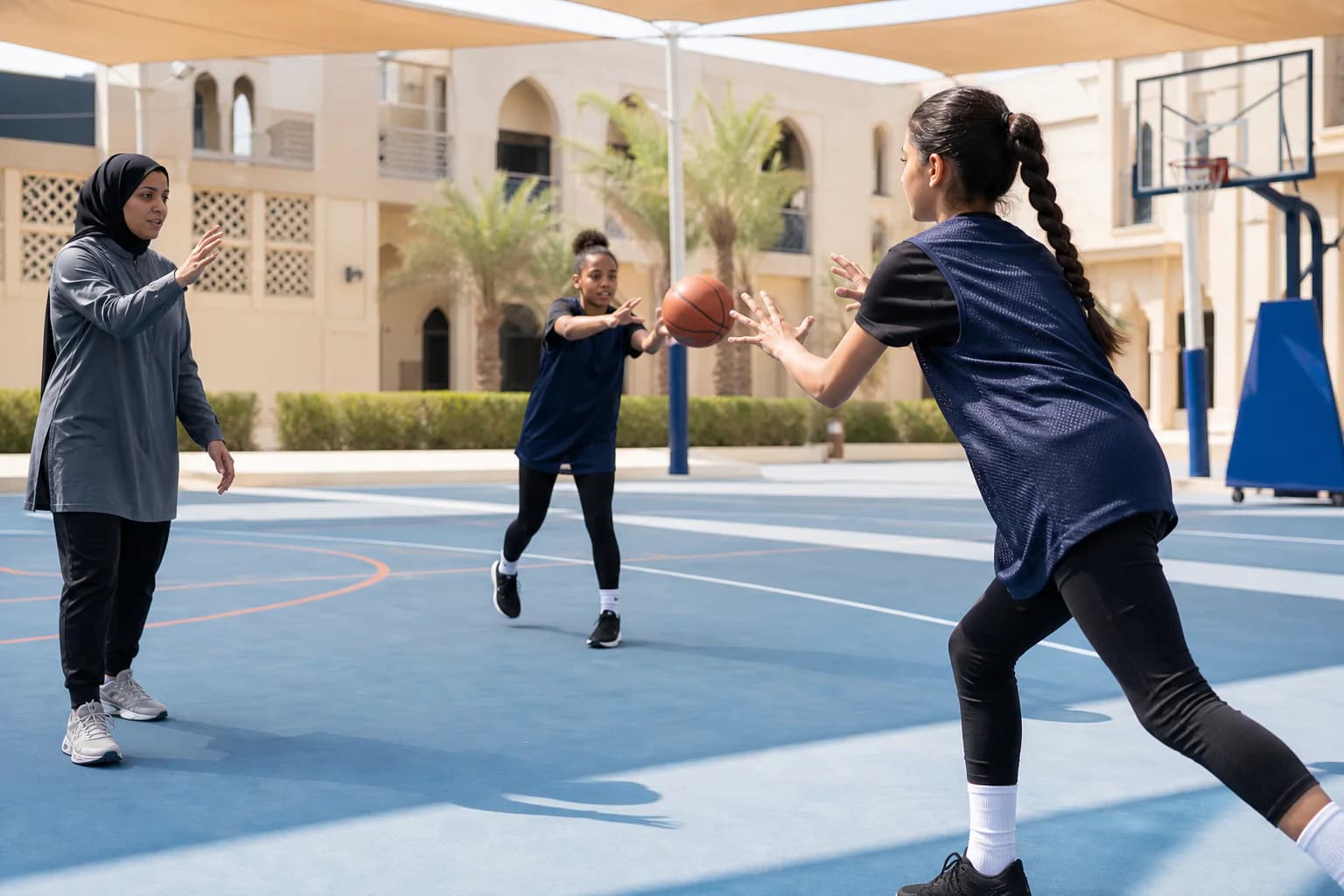 Girls practising accurate basketball passing with a coach on a shaded Dubai court